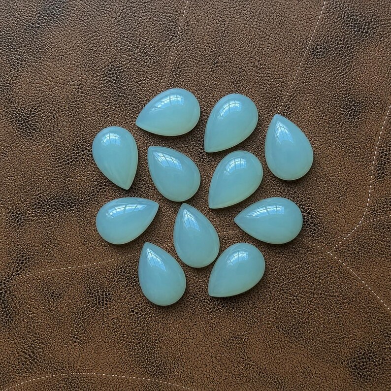 Pu&ograve; includere: Una collezione di dieci gemme a forma di goccia di colore verde pallido. Le pietre lucide sono disposte su una superficie marrone strutturata, ognuna con una parte superiore liscia e arrotondata e una base appuntita. Le gemme sono traslucide.
