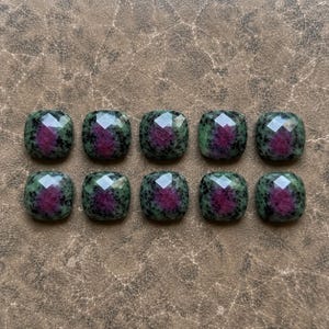 May include: Ten square-shaped, faceted zoisite gemstones arranged in two rows of five. Each stone features a deep magenta center surrounded by a mottled green and black pattern, with a polished, reflective surface showing geometric facets.