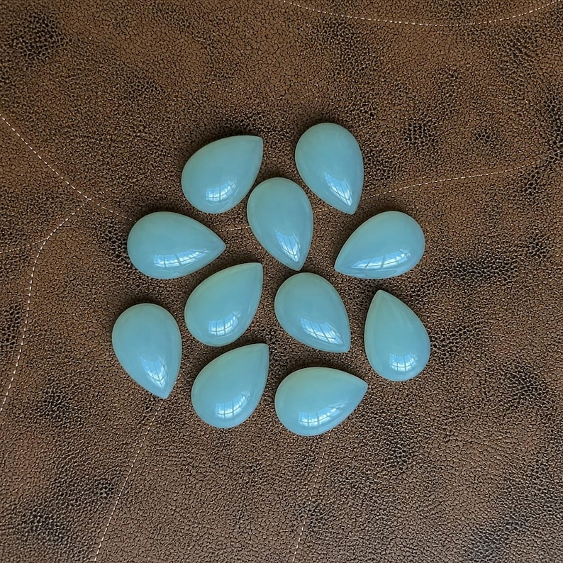 Pu&ograve; includere: Una collezione di dieci gemme a forma di goccia di colore azzurro pallido. Le pietre lucide sono disposte su una superficie marrone simile alla pelle. Le gemme hanno una finitura liscia e lucida e riflettono la luce.