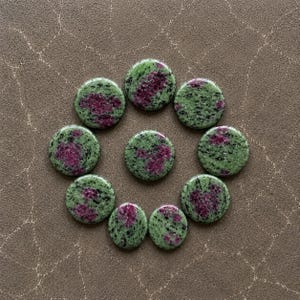 Puede incluir: Diez cabujones circulares de zoisita con inclusiones de rubí dispuestos en un patrón circular. Cada piedra presenta una base verde claro moteada con puntos negros y parches irregulares distintivos de mineral de rubí color magenta intenso.