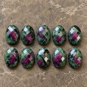 Könnte beinhalten: Zehn oval geformte Cabochons, angeordnet in zwei Reihen zu je fünf Stück. Jeder Cabochon hat eine facettierte Oberfläche mit einer Mischung aus dunkelgrünen und schwarzen Sprenkelmustern, akzentuiert durch leuchtend magentafarbene Einschlüsse.