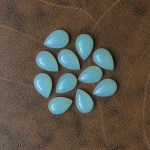 Pu&ograve; includere: Una collezione di undici gemme a forma di goccia di colore verde pallido. Le pietre lucide sono disposte su una superficie marrone simile alla pelle. Ogni pietra ha una finitura liscia e lucida, che riflette la luce. Le gemme sono lunghe circa 2 cm.