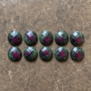 May include: Ten round, faceted cabochons made of zoisite with ruby inclusions. Each stone features a dark green and black speckled base with a vibrant, deep pinkish-red center. The stones are arranged in two horizontal rows of five pieces each.
