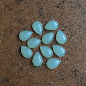Pu&ograve; includere: Una collezione di undici gemme a forma di goccia di colore verde pallido. Le pietre lucide sono disposte su una superficie marrone simile alla pelle. Le gemme hanno una finitura liscia e lucida e sono illuminate in modo uniforme.