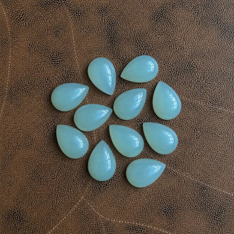 Pu&ograve; includere: Una collezione di undici gemme a forma di goccia di colore verde pallido. Le pietre lucide sono disposte su una superficie marrone simile alla pelle. Le gemme hanno una finitura liscia e lucida e sono illuminate in modo uniforme.