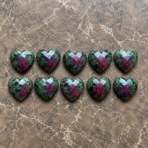 May include: Ten faceted heart-shaped stones arranged in two rows of five. Each stone features a mottled green and black surface with a prominent, vibrant magenta-pink center, showcasing a polished, reflective finish on the top facets.
