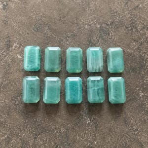May include: Ten rectangular, light teal gemstones arranged in two rows of five. Each stone has a faceted surface, catching the light. The stones are a consistent size and shape, with a slight variation in internal patterns. The background is a mottled, neutral color.