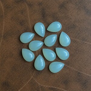 Pu&ograve; includere: Una collezione di undici gemme a forma di goccia di colore verde pallido. Le pietre lucide sono disposte su una superficie marrone simile alla pelle. Le gemme hanno una finitura liscia e lucida.