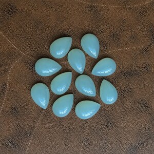 Pu&ograve; includere: Una collezione di dieci gemme a forma di goccia di colore azzurro pallido. Le pietre lucide sono disposte su una superficie marrone simile alla pelle. Le gemme hanno una finitura liscia e lucida e riflettono la luce.