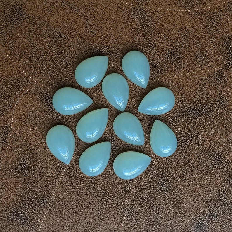 Pu&ograve; includere: Una collezione di dieci gemme a forma di goccia di colore azzurro pallido. Le pietre lucide sono disposte su una superficie marrone simile alla pelle. Le gemme hanno una finitura liscia e lucida e riflettono la luce.