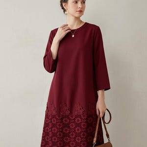 Può includere: Un abito color bordeaux scuro al ginocchio con maniche a tre quarti, scollo rotondo e un orlo in pizzo decorativo con motivo floreale. La parte inferiore dell'abito è rifinita con un'ampia fascia di pizzo rosso scuro abbinato.