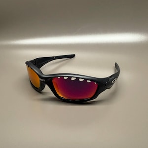May include: Black sunglasses with red-orange mirrored lenses. The sunglasses have a curved frame and the word "SATISFY" is printed on the left lens. The lenses are a vibrant red-orange color.