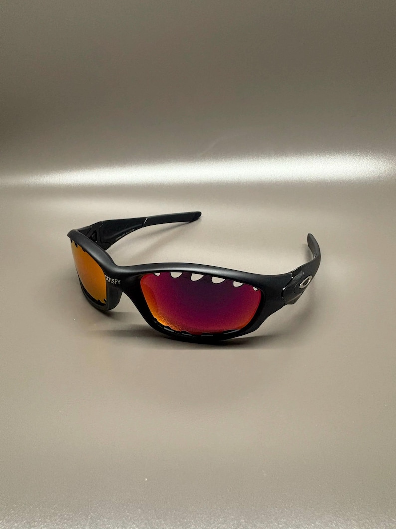 May include: Black sunglasses with red-orange mirrored lenses. The sunglasses have a curved frame and the word "SATISFY" is printed on the left lens. The lenses are a vibrant red-orange color.