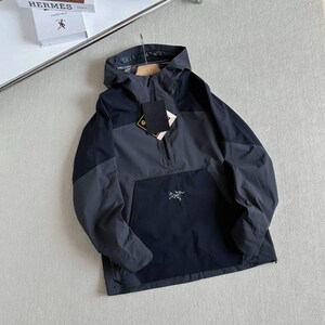 May include: A dark gray and navy blue anorak jacket with a hood and a half-zip closure. The jacket features a front pocket with a small logo and a hang tag. The jacket is hanging on a wooden hanger.
