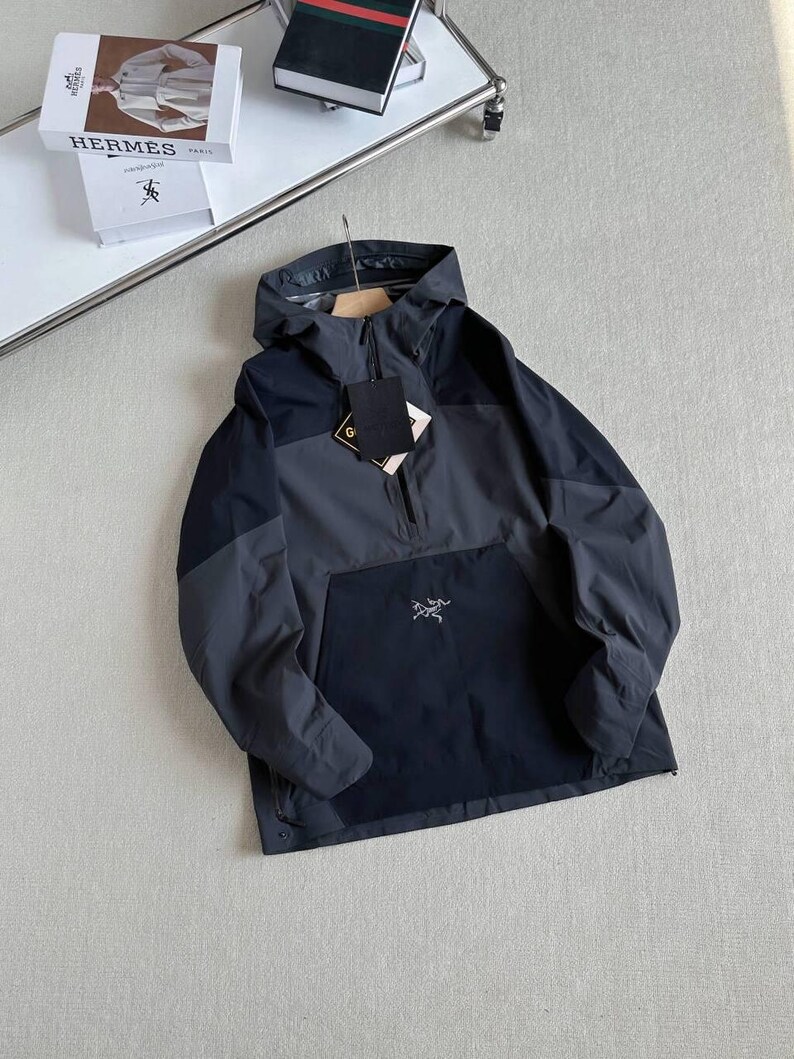 May include: A dark gray and navy blue anorak jacket with a hood and a half-zip closure. The jacket features a front pocket with a small logo and a hang tag. The jacket is hanging on a wooden hanger.