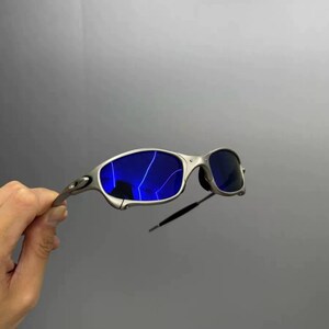 Może przedstawiać: Okulary przeciwsłoneczne w srebrnej oprawie z jaskrawoniebieskimi soczewkami. Okulary mają elegancki, nowoczesny design z zakrzywioną oprawą i ciemnymi zausznikami. Soczewki odbijają światło.