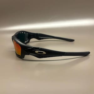 May include: Black sunglasses with orange-tinted lenses. The sunglasses have a sleek, modern design with a black frame and arms. The lenses are a vibrant orange color, and the overall look is sporty and stylish.