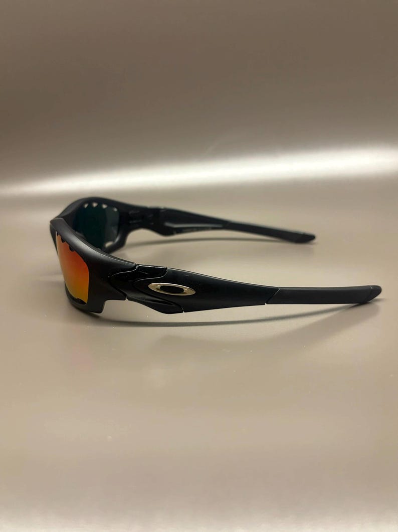 May include: Black sunglasses with orange-tinted lenses. The sunglasses have a sleek, modern design with a black frame and arms. The lenses are a vibrant orange color, and the overall look is sporty and stylish.
