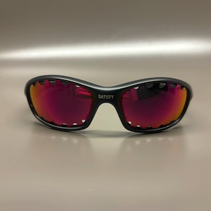 May include: Black sunglasses with red-tinted lenses. The sunglasses have a curved frame with a unique scalloped edge design. The word "SATISFY" is printed on the bridge of the sunglasses.
