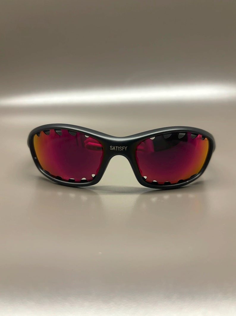 May include: Black sunglasses with red-tinted lenses. The sunglasses have a curved frame with a unique scalloped edge design. The word "SATISFY" is printed on the bridge of the sunglasses.