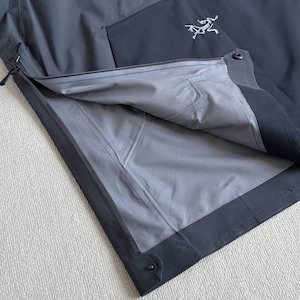May include: A dark gray and black jacket with a partial zipper and a white logo on the chest. The jacket's interior is a lighter gray, and the seams are visible. The jacket is made of a waterproof material.