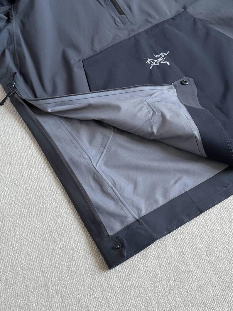 May include: A dark gray and black jacket with a partial zipper and a white logo on the chest. The jacket's interior is a lighter gray, and the seams are visible. The jacket is made of a waterproof material.
