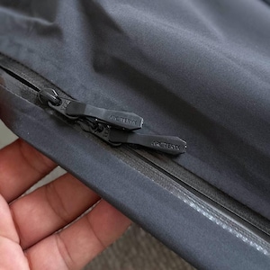 May include: Close-up of a dark gray jacket with a black zipper. The zipper pull features the "ARC'TERYX" logo. The jacket material appears smooth and water-resistant, with a subtle sheen. The image highlights the jacket's construction and branding.