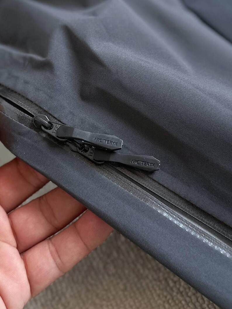 May include: Close-up of a dark gray jacket with a black zipper. The zipper pull features the "ARC'TERYX" logo. The jacket material appears smooth and water-resistant, with a subtle sheen. The image highlights the jacket's construction and branding.