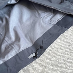 May include: Close-up of a gray jacket interior. The jacket has a light gray lining and a dark gray exterior with a drawstring and a small, rectangular fabric tag. The jacket appears to be made of a waterproof material.
