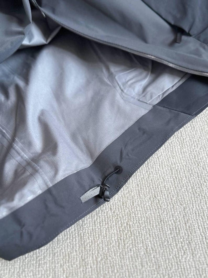 May include: Close-up of a gray jacket interior. The jacket has a light gray lining and a dark gray exterior with a drawstring and a small, rectangular fabric tag. The jacket appears to be made of a waterproof material.