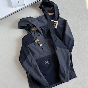 May include: Two hooded jackets in shades of gray and black are displayed. The jackets have a half-zip closure and a small logo on the front. Each jacket has a tag attached. The jackets are on a light-colored surface.