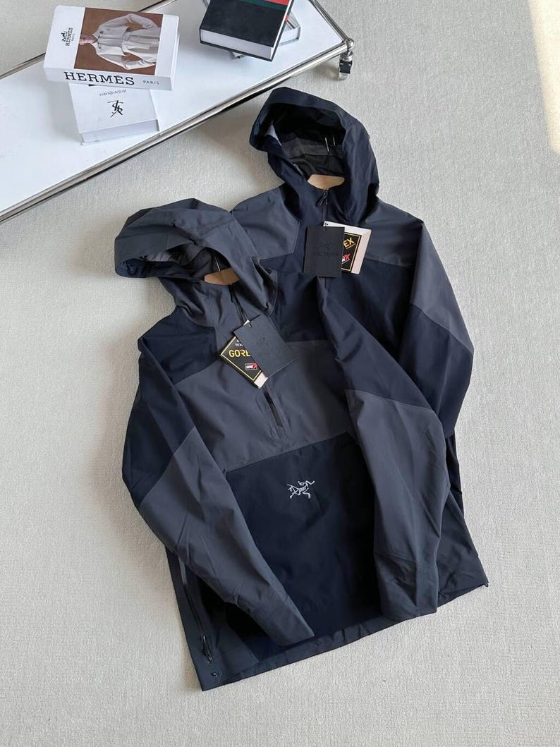 May include: Two hooded jackets in shades of gray and black are displayed. The jackets have a half-zip closure and a small logo on the front. Each jacket has a tag attached. The jackets are on a light-colored surface.