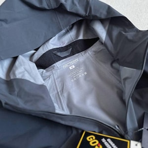 May include: A dark gray hooded jacket with the brand name "Arc'teryx" visible on the inside lining. The jacket has a tag that reads "GORE-TEX" and includes a guarantee to keep you dry. The jacket is made of a waterproof material.
