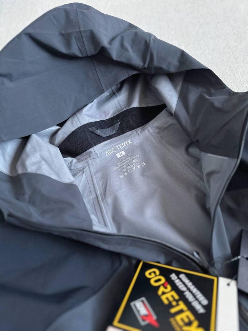 May include: A dark gray hooded jacket with the brand name "Arc'teryx" visible on the inside lining. The jacket has a tag that reads "GORE-TEX" and includes a guarantee to keep you dry. The jacket is made of a waterproof material.