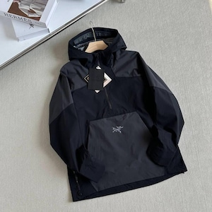 May include: A black and gray hooded anorak jacket with a front kangaroo pocket and a half-zip closure. The jacket features a small logo on the pocket and a tag with text. The jacket is hanging on a wooden hanger.