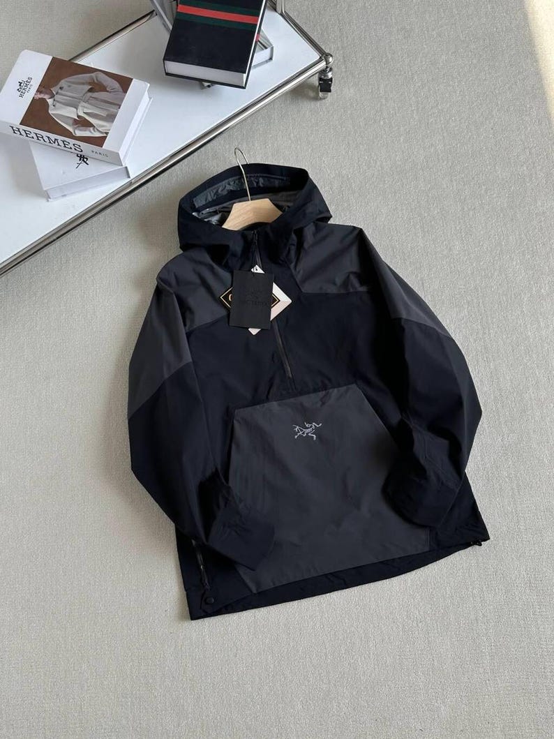 May include: A black and gray hooded anorak jacket with a front kangaroo pocket and a half-zip closure. The jacket features a small logo on the pocket and a tag with text. The jacket is hanging on a wooden hanger.