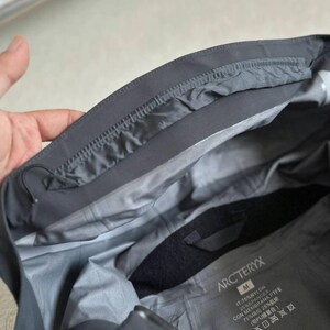 May include: A dark gray jacket interior with a label that reads "ARC'TERYX". The jacket is made of nylon and spandex. The inside of the jacket is a lighter gray color.