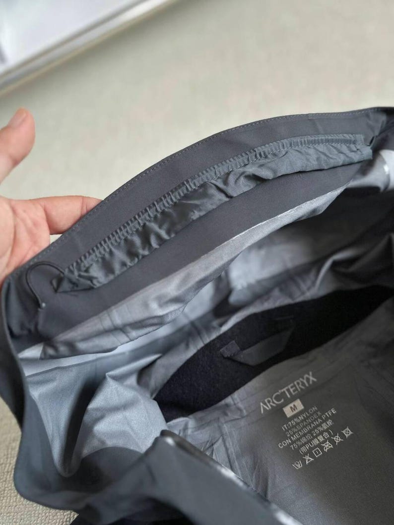 May include: A dark gray jacket interior with a label that reads "ARC'TERYX". The jacket is made of nylon and spandex. The inside of the jacket is a lighter gray color.
