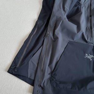 May include: Two dark gray and black jackets are layered on a light gray surface. The jackets have multiple zippered pockets and a drawstring at the hem. The Arc'teryx logo is visible on the right jacket.