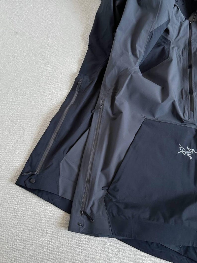 May include: Two dark gray and black jackets are layered on a light gray surface. The jackets have multiple zippered pockets and a drawstring at the hem. The Arc'teryx logo is visible on the right jacket.