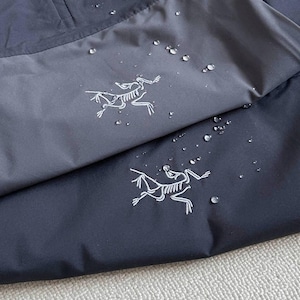 May include: Close-up of a navy blue and gray jacket with a zipper and a white logo of a running creature. Water droplets are visible on the fabric, suggesting a waterproof design. The jacket appears to be made of a technical fabric.