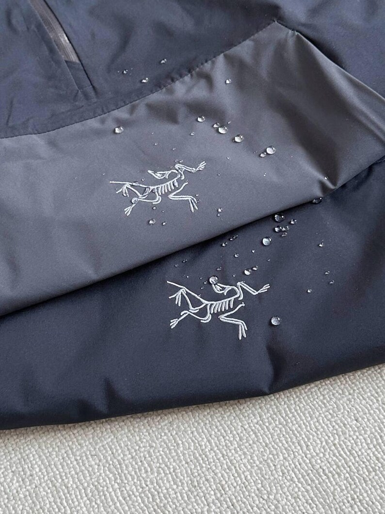 May include: Close-up of a navy blue and gray jacket with a zipper and a white logo of a running creature. Water droplets are visible on the fabric, suggesting a waterproof design. The jacket appears to be made of a technical fabric.