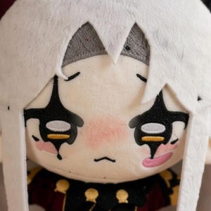 May include: A soft plush doll featuring a white head covering and a detailed face. The doll has black eye markings, pink cheeks, and a stitched mouth. The doll's outfit includes a dark red and gold patterned top.