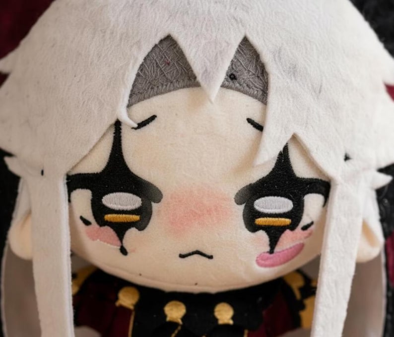 May include: A soft plush doll featuring a white head covering and a detailed face. The doll has black eye markings, pink cheeks, and a stitched mouth. The doll's outfit includes a dark red and gold patterned top.