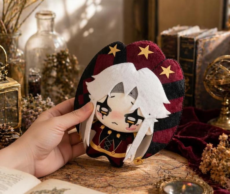 May include: A plush doll with white hair and a red and black outfit. The doll has a sad expression and is held in a hand. The background includes books and decorative items.