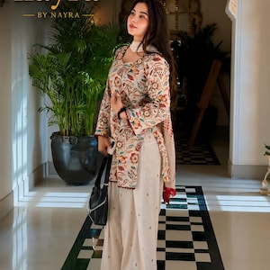Op de afbeelding: Een beige tuniek met lange mouwen en een veelkleurige bloemenprint, gecombineerd met een wijde, gebroken witte broek met kleine geborduurde details. De outfit bevat een bijpassende dupatta en de tekst label nayra BY NAYRA.