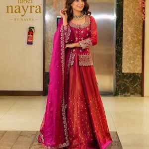 May include: A vibrant magenta and deep red ensemble featuring a long-sleeved jacket with intricate silver embroidery and mirror work, paired with a matching floor-length skirt and a sheer dupatta with a decorative border. Text reads label nayra.