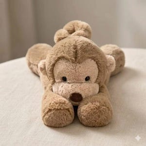 May include: A plush, light brown monkey toy with a soft, furry texture. The monkey is lying down with its arms and legs outstretched. It has a round face, small black eyes, and a brown nose. The toy is designed to be cuddly.