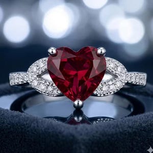 Peut inclure: Une bague argentée ornée d'une grande pierre précieuse rouge foncé en forme de cœur au centre. L'anneau se divise en deux bras incurvés délicats de chaque côté, incrustés de petites pierres claires et scintillantes le long de la tige.