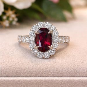 Puede incluir: Un anillo con una gran piedra preciosa de color rojo intenso y corte ovalado, montada en una banda de metal plateado. La piedra central está rodeada por un halo de pequeñas piedras claras y brillantes, con más piedras en la banda.